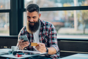 A bearded man manages his credit card from his phone at a cafe table.